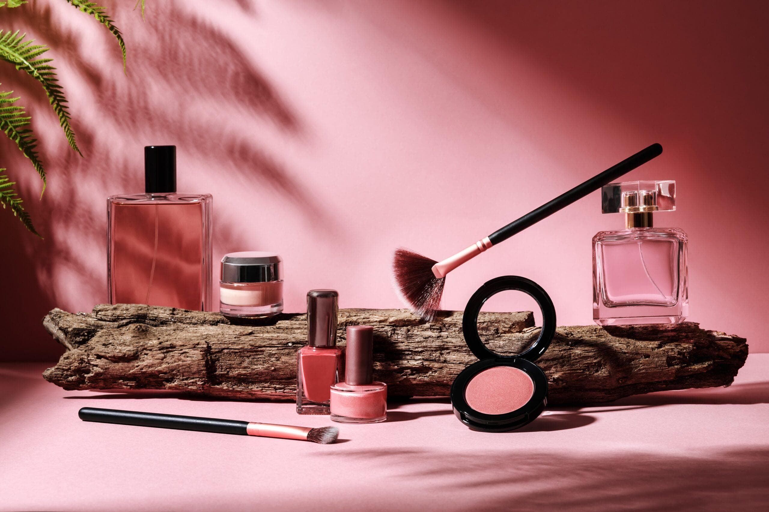 Perfume, nail polish, and blush styled on driftwood against a pink backdrop